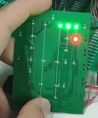 A prototype circuit board held in the hand with several LEDs lit, connected by wires to a breadboard on a workbench.