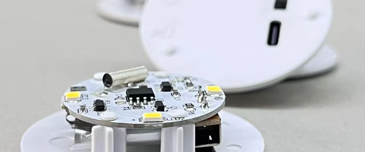 A round white PCB with four LEDs, a driver IC, a tactile switch and a USB-C socket, resting on white housing bases.